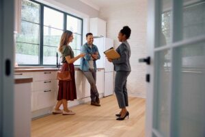 A young couple with a notepad tours a bright, unfurnished home with hardwood floors and exposed brick walls.