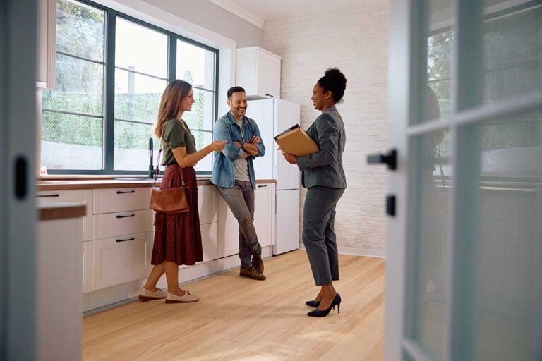A young couple with a notepad tours a bright, unfurnished home with hardwood floors and exposed brick walls.